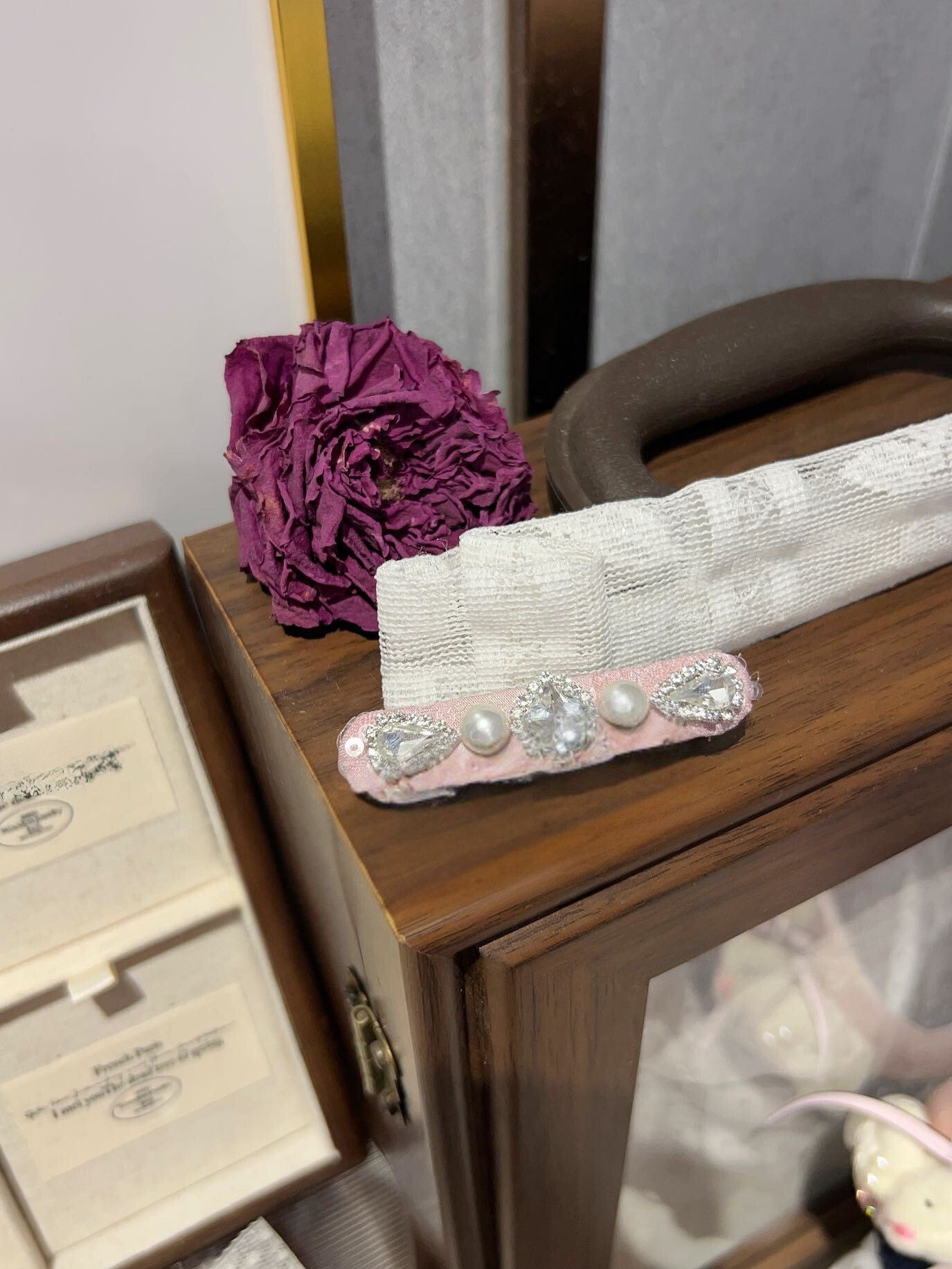 Decorative lace hair clip with pearls and Swarovski crystals on a wooden surface next to a purple flower and jewelry box. Only at Petite Murmure