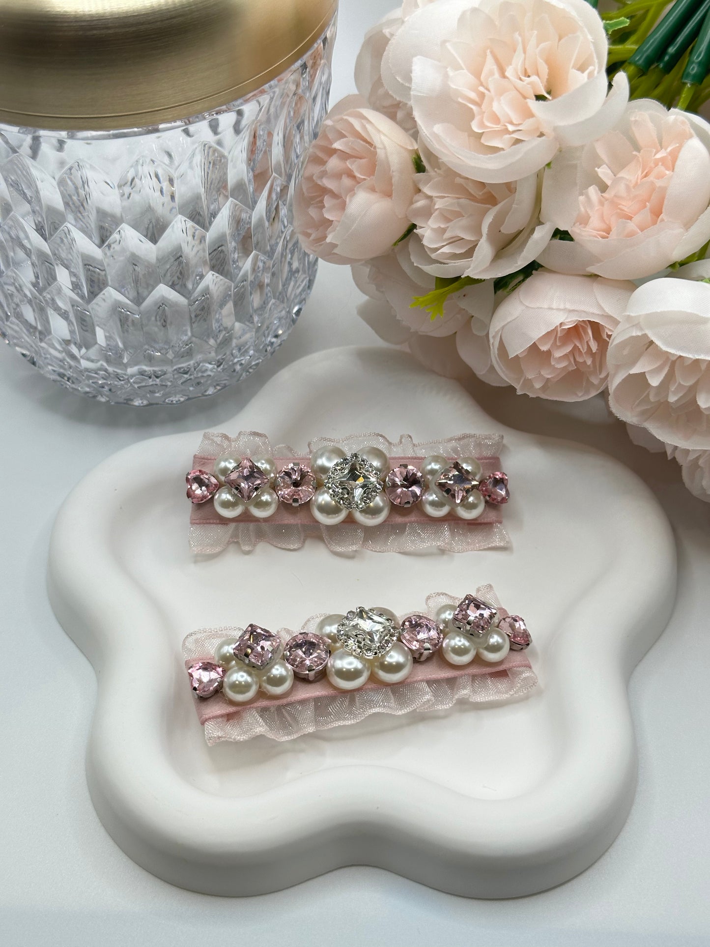 Pair of hair clips with Swarovski crystals and pearls embellished and silky organza on white star-shaped plate, surrounded by flowers and a crystal vase. Only at petite murmure