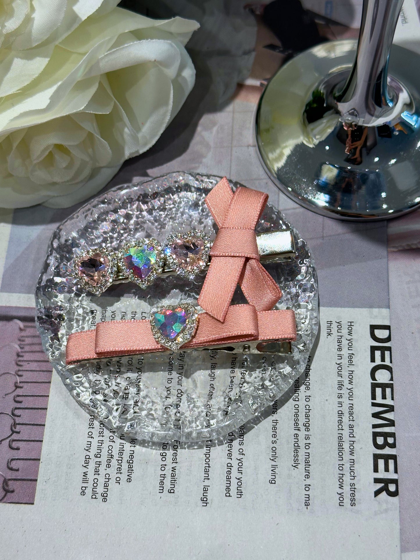 Set of Two pink decorative hair clips with pink bows and heart shape Swarovski crystal  details on a round, glittery base with white roses and a candle in the background.