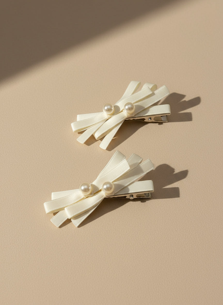 Two cream white bow hair clips with layered ribbons and pearl centers on a beige background