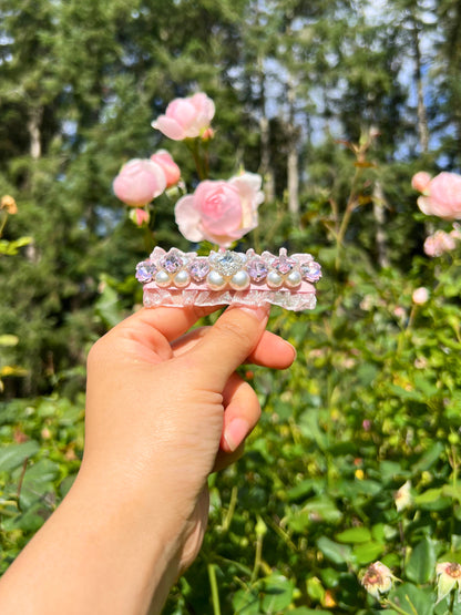 Hand holding a hair clip with Swarovski crystals and pearls embellished and silky organza  with pink flowers in the background. Only at Petite Murmure