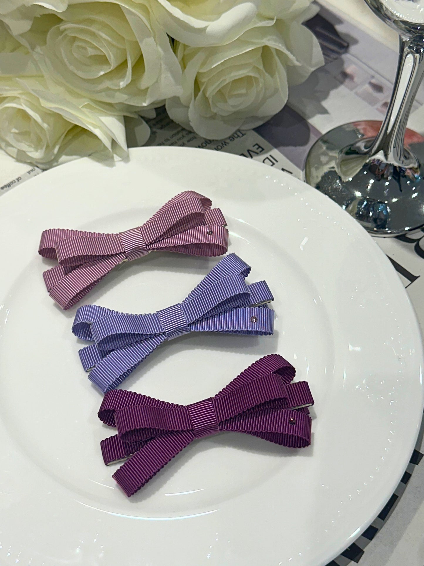 Three purple hair bows on a white plate with flowers and a glass in the background.