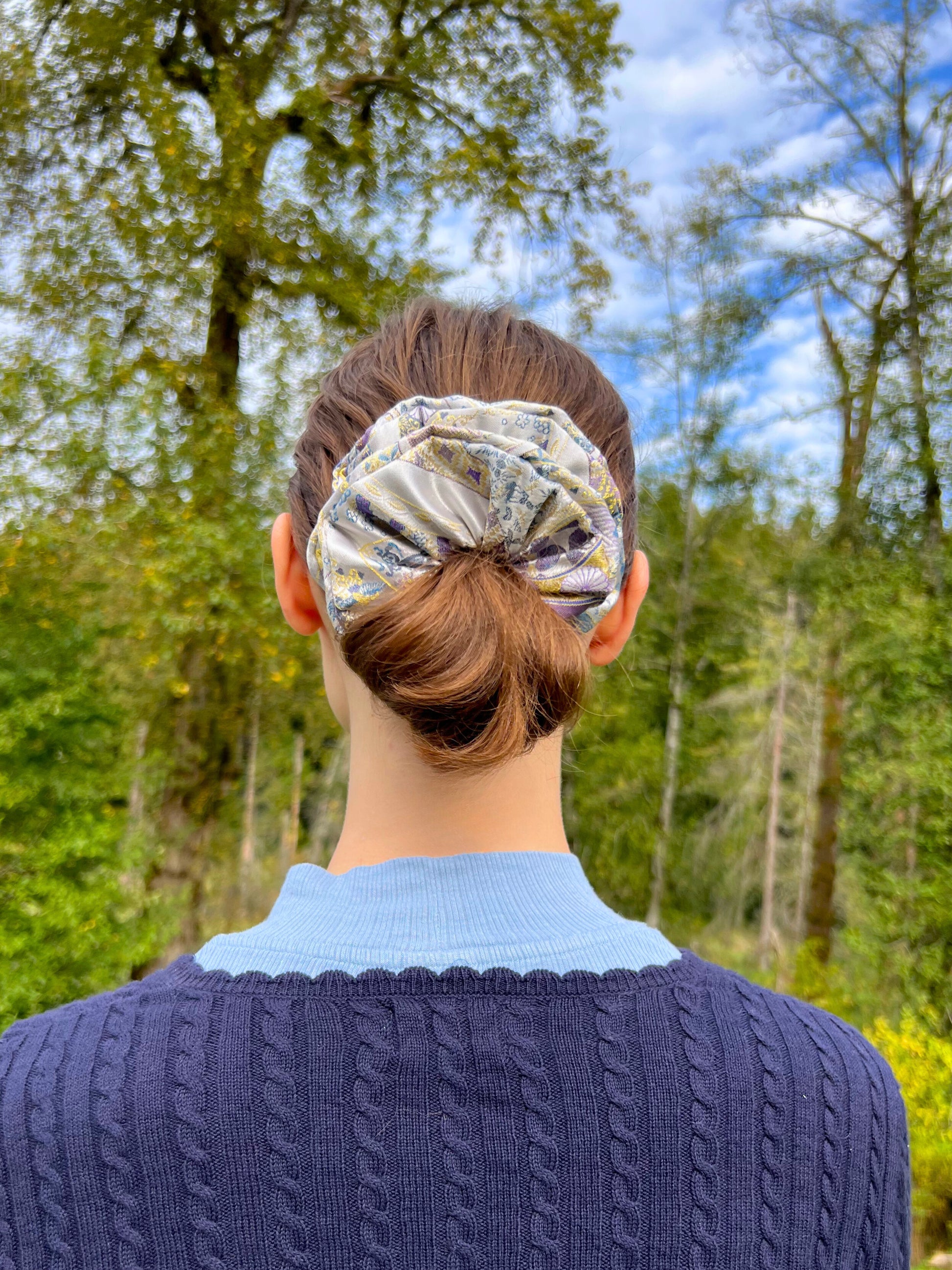 Person wearing a Exclusive Song brocade mulberry silk scrunchie with peony aromatic scent, infused with gold thread. Phoenix patterned scrunchie outdoors with trees and blue sky in the background