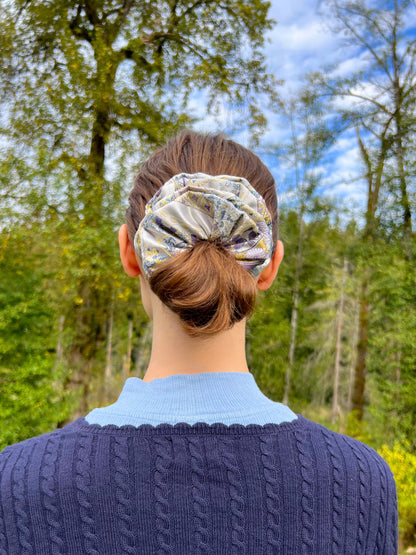 Person wearing a Exclusive Song brocade mulberry silk scrunchie with peony aromatic scent, infused with gold thread. Phoenix patterned scrunchie outdoors with trees and blue sky in the background
