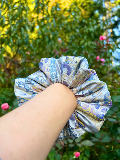 Hand holding a Exclusive Song brocade mulberry silk scrunchie with peony aromatic scent, infused with gold thread. Phoenix patterned scrunchie with a blurred green outdoor background