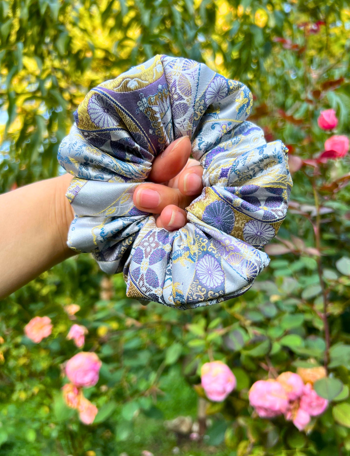 Hand holding the Exclusive Song brocade mulberry silk scrunchie with peony aromatic scent, infused with gold thread. Phoenix patterned scrunchie with a garden background