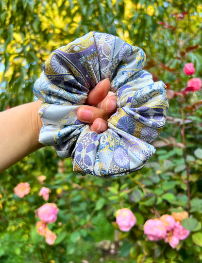 Hand holding the Exclusive Song brocade mulberry silk scrunchie with peony aromatic scent, infused with gold thread. Phoenix patterned scrunchie with a garden background
