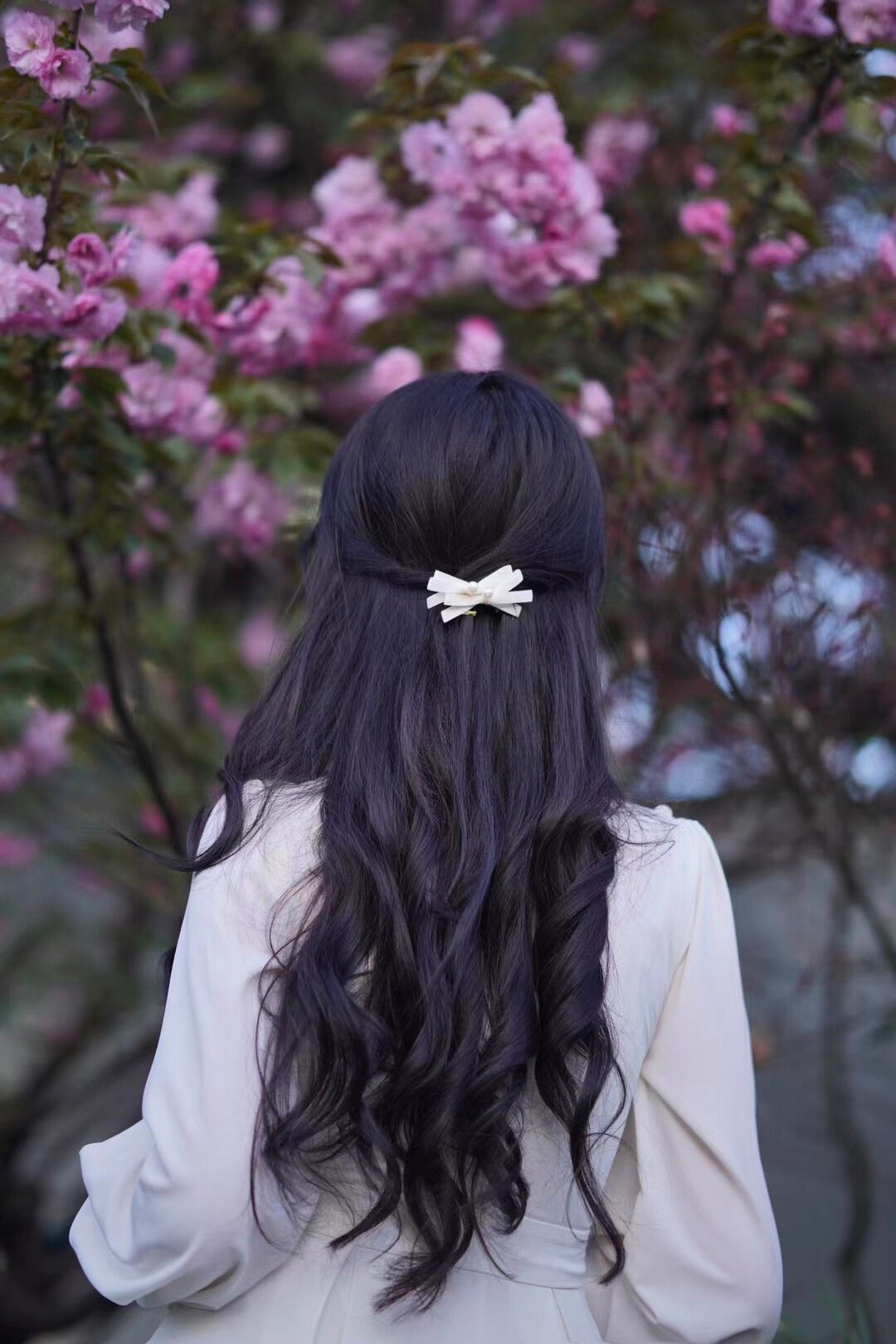 Person with long dark hair and a creamy white bow standing in front of pink cherry blossoms.