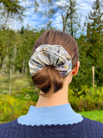 Person wearing an Exclusive Song brocade mulberry silk scrunchie with peony aromatic scent, infused with gold thread. Phoenix patterned scrunchie outdoors with trees and sky in the background