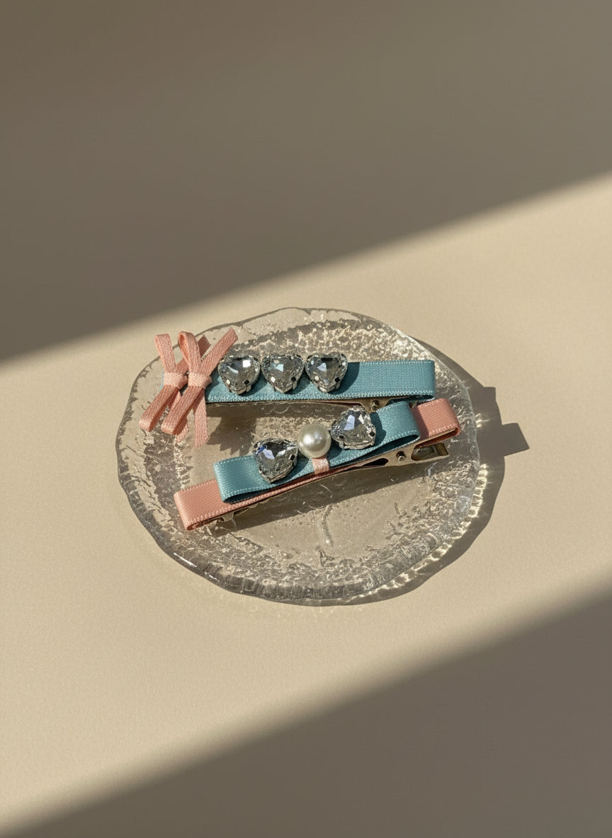 Set of Two decorative hair clips with pink and blue ribbons and pearl and Swarovski crystals on a clear glass plate against a beige background