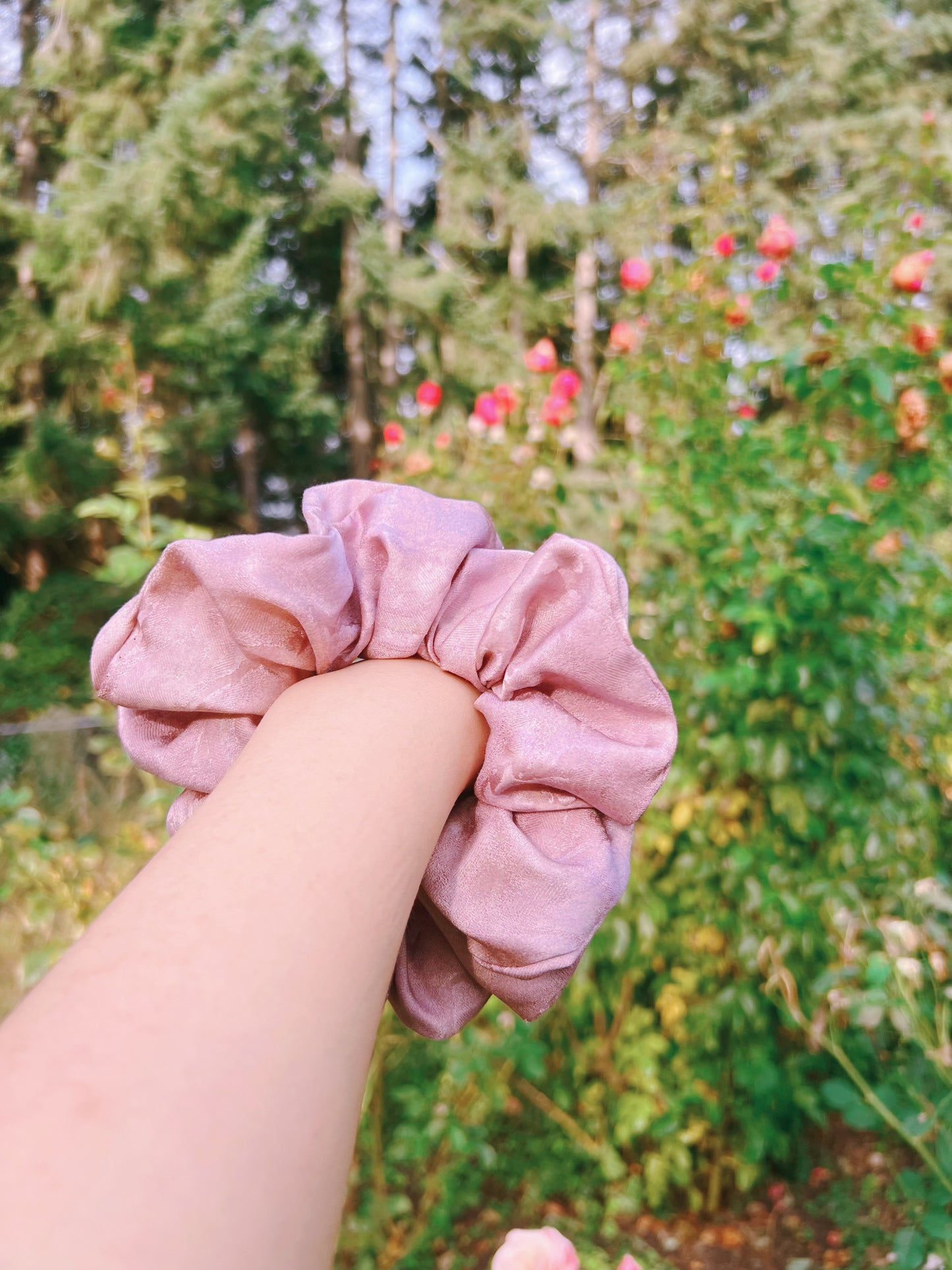 Pink handmade rose aromatic scented mulberry silk scrunchie held by a hand with a blurred green and pink floral background