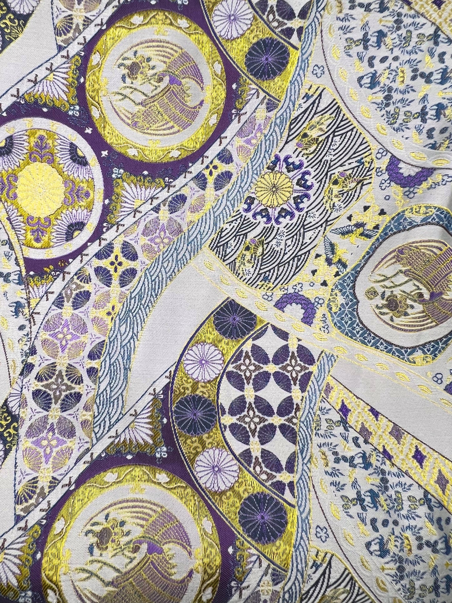 Close-up of a patterned fabric with circular designs in purple, yellow, and white to make the Exclusive Song brocade mulberry silk scrunchie with peony aromatic scent, infused with gold thread. Phoenix patterned scrunchie